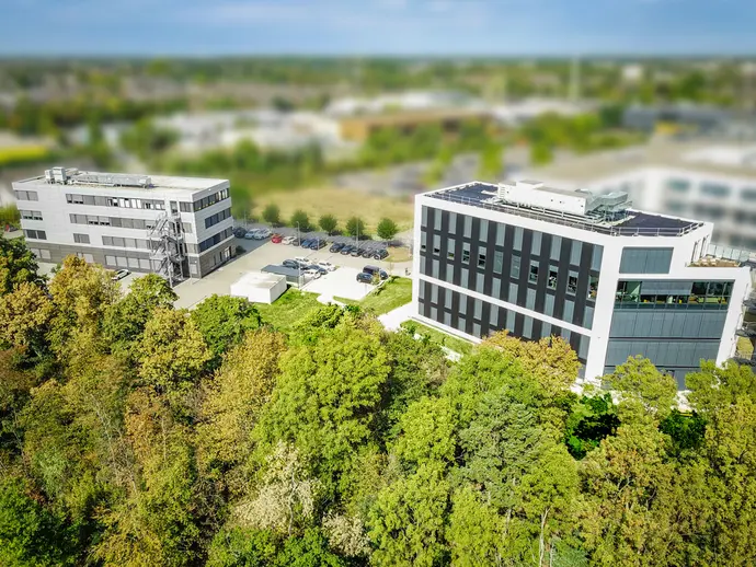 Aerial view of two modern buildings surrounded by trees. The left building is gray with a staircase, while the right building features a sleek black facade with large windows, and solar panels on the roof. A parking lot with several cars is visible nearby.