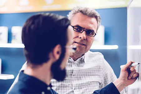 Two men engage in a discussion, with one man writing on a whiteboard. The man writing appears focused and is using a marker, while the other man listens attentively, maintaining eye contact, suggesting an interactive learning or brainstorming session.