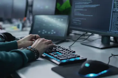 A pair of hands typing on a backlit keyboard, with two computer monitors displaying coding and software interfaces in the background. A computer mouse is positioned nearby. The environment appears focused on programming or tech work.
