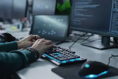 A pair of hands typing on a backlit keyboard, with two computer monitors displaying coding and software interfaces in the background. A computer mouse is positioned nearby. The environment appears focused on programming or tech work.