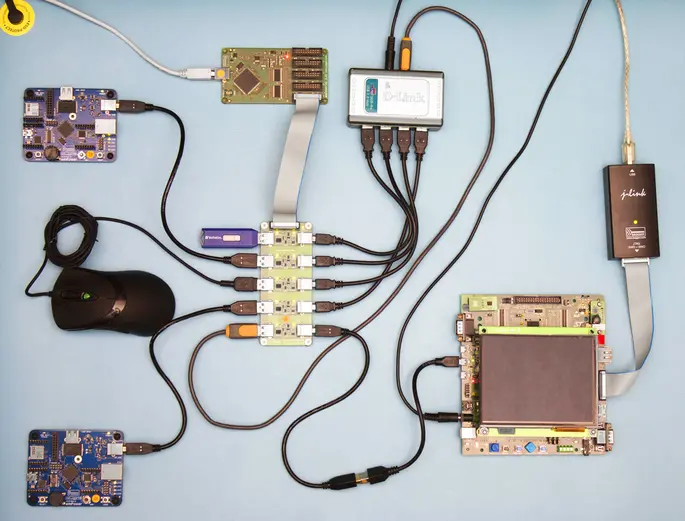 The image shows a collection of circuit boards and electronic components interconnected with various cables. A mouse and USB devices are also present, illustrating a setup likely used for testing or developing electronics. The background is a solid light color, emphasizing the hardware.