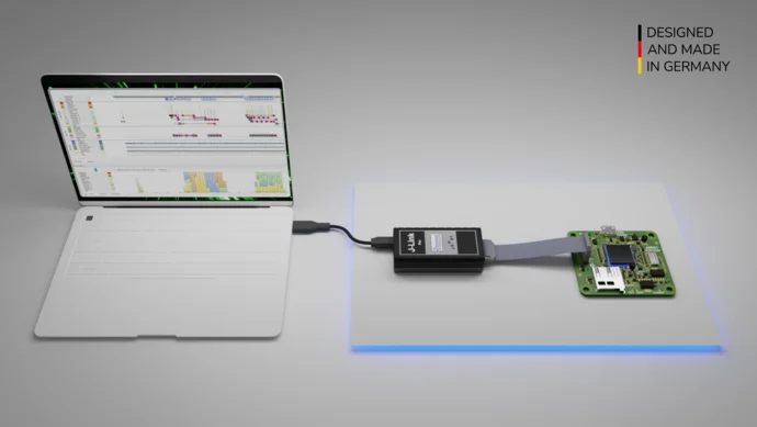 An open laptop displays software on its screen, connected via a cable to a black device labeled "J-Link." The device links to a green circuit board with various components and connectors, set against a softly illuminated background. The text in the corner highlights "DESIGNED AND MADE IN GERMANY.