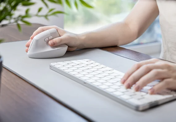 A person's hand is holding a white computer mouse while the other hand is typing on a sleek, white keyboard. The setup is on a gray desk surface, with a blurred background suggesting a bright, indoor environment.