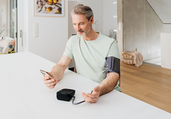 A middle-aged man sits at a table, checking his smartphone while wearing a blood pressure monitor on his arm. A black blood pressure device rests on the table in front of him. The setting is bright and modern.