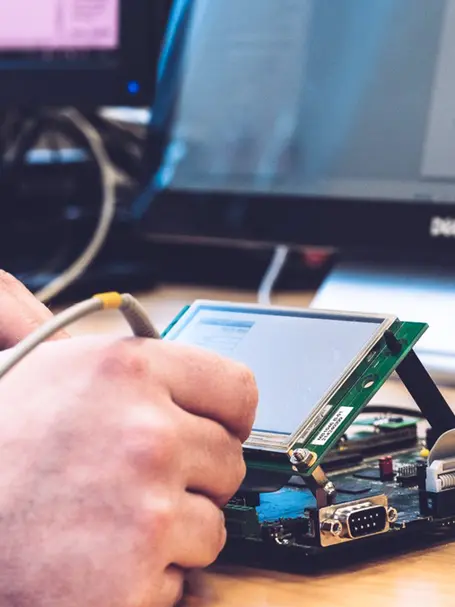 A hand is seen connecting a wire to a circuit board with a small display screen. The setup is likely related to electronics or programming, with components arranged on a table. In the background, a computer monitor is partially visible.