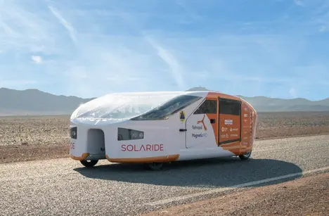 A futuristic solar-powered vehicle named "Solaride" is parked on a road in a barren landscape with mountains in the background. The vehicle features a sleek, aerodynamic design and is predominantly white with orange accents and branding.