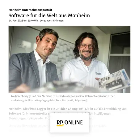 Two men are smiling in an office environment. They are identified as Ivo Geilenbruegge and Dirk Akemann, showcasing their pride in their company culture. In the background, there is a chart or display likely related to technology. The image emphasizes their collaboration in software development for microcontrollers.