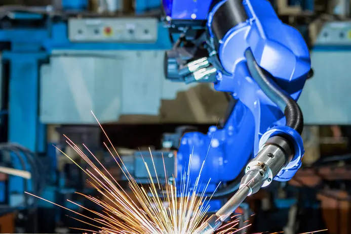 A blue robotic arm is positioned to weld, emitting bright sparks. The background features machinery, suggesting an industrial environment. The scene captures the action of automated manufacturing.