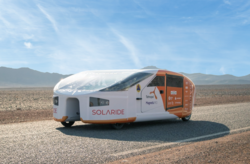 A futuristic solar-powered vehicle named "Solaride" is parked on a desert road. The vehicle has a sleek, aerodynamic design with a white and orange color scheme, and various logos displayed on its sides. Mountains are visible in the background under a clear blue sky.
