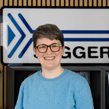 A smiling person with short gray hair and glasses stands in front of a backdrop featuring a large sign with blue arrows and the word "JIGGER." The setting has wooden paneling and items displayed on a shelf behind them.