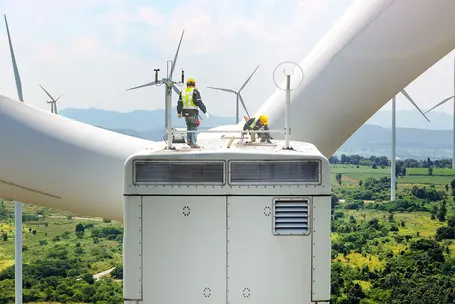 Two workers in safety gear are performing maintenance on top of a large wind turbine. The scene includes other wind turbines in the background against a blue sky and green landscape, emphasizing the renewable energy environment.