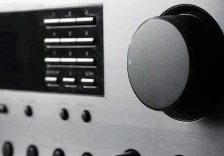 Close-up view of a silver audio equipment panel featuring buttons and a prominent round volume knob. The knob is black and has a subtle dot indicating its position. The background texture is sleek and metallic, with various other control elements visible.