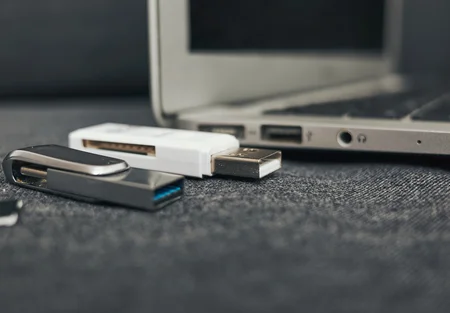 The image shows two USB flash drives, one gray and one white, placed on a textured surface. A laptop is visible in the background, highlighting its USB ports. The setting conveys a theme of digital storage and technology.