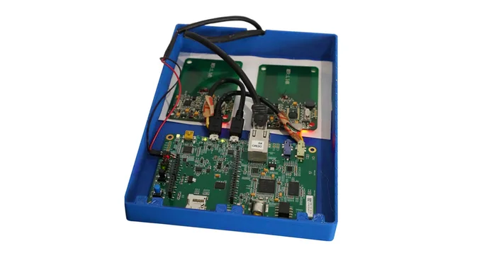 An electronic circuit board is mounted inside a blue casing. It features various components, including connectors and a lit indicator. Additional smaller boards are placed on top, showcasing circuitry and components for electronic functions. Cables are visible, connecting the parts together.