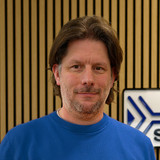 A man with medium-length dark hair and a beard is smiling, wearing a blue sweater. He stands in front of a wooden wall with vertical stripes and a sign that reads "SEGGER." The setting appears to be a modern office or workspace.