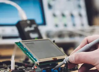A hand is holding a probe, testing connections on a circuit board with a small screen attached. The background includes electronic equipment, suggesting a workspace focused on electronics and diagnostics.