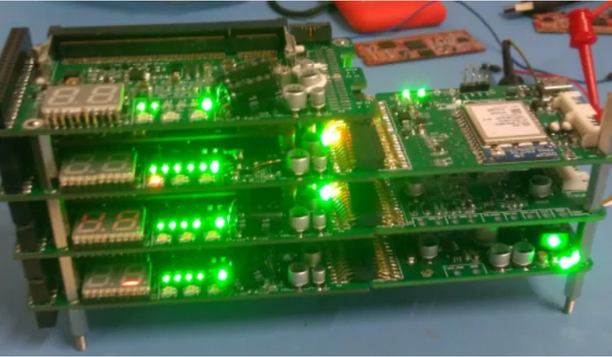 A stack of green-lit circuit boards is displayed on a blue surface. The boards feature various components, including digital displays showing numbers. Some boards have a mix of green and orange lights, indicating active circuits and functionality within the assembled device.