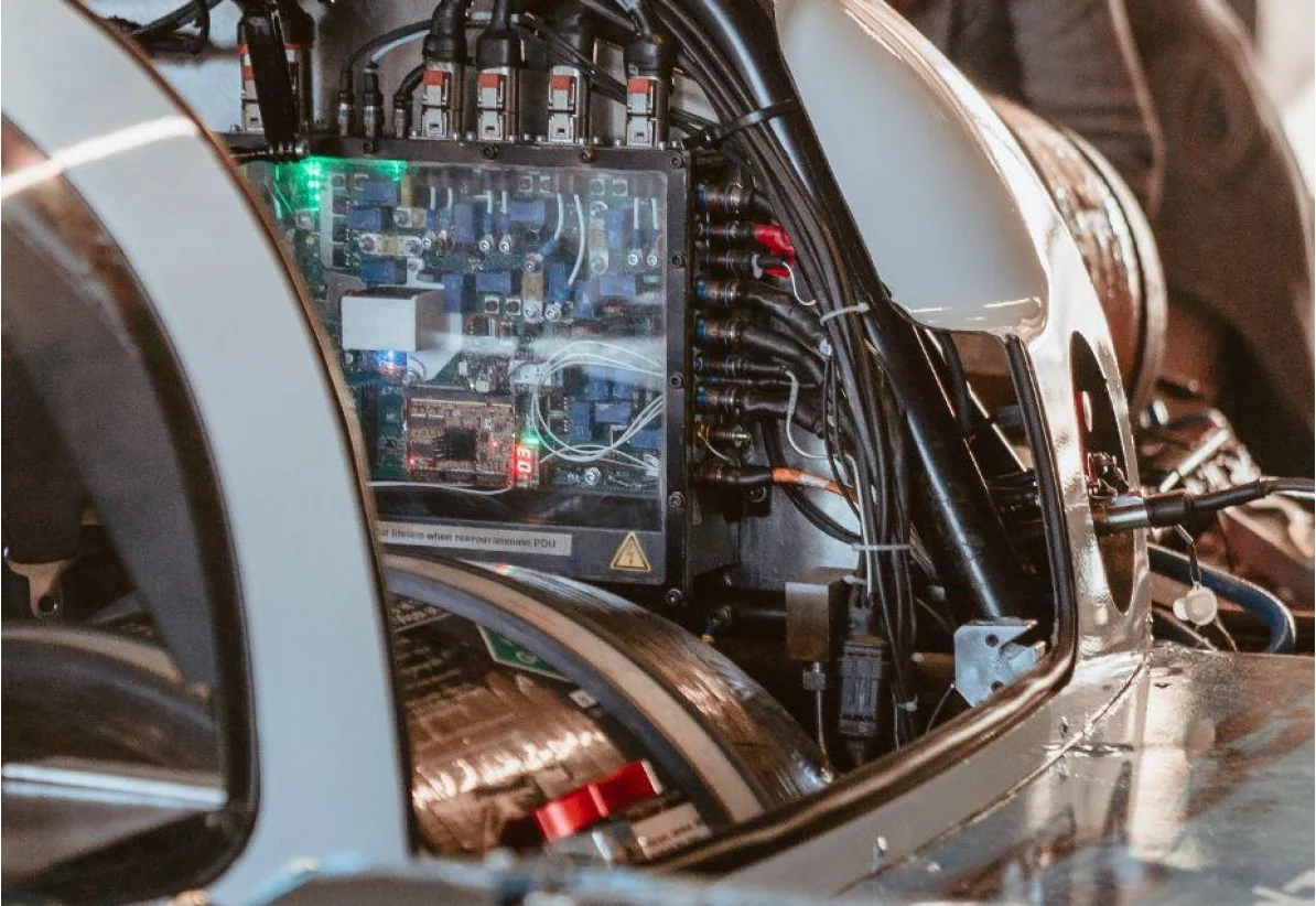 The image shows the interior of a vehicle, highlighting a complex arrangement of electronic components, wiring, and control panels. Visible elements include a clear panel displaying circuit boards and indicator lights, surrounded by various cables and connectors integral to the vehicle's systems.