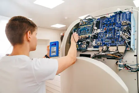 A technician inspects and repairs a device's internal circuitry, holding a diagnostic tool in one hand. The device features a complex arrangement of blue circuit boards and wires, indicating a technical and possibly medical setting. Bright overhead lighting illuminates the workspace.