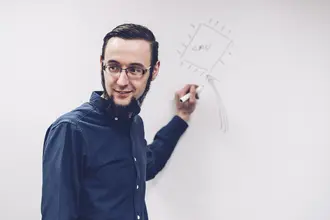 A person with glasses and a beard is standing beside a whiteboard, smiling as they use a marker to draw a rectangle labeled "CPU" with arrows indicating connections. The setting suggests a teaching or brainstorming environment.