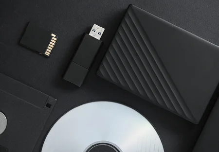A flat lay of various data storage devices is displayed on a black background. Items include an SD card, a USB flash drive, an external hard drive with a ribbed design, a floppy disk, and a CD.