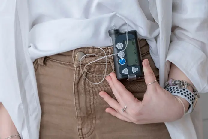 A person wearing a white shirt and brown pants is holding an insulin pump attached to their waistband. The pump has a digital display and buttons, with a small tube connected to it. The person's hand is resting near the device, indicating it is in use.