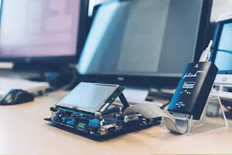 The image shows an electronic development board with a connected display screen and a communication device on a workspace. Computer monitors are in the background, highlighting a tech-focused environment for electronics and programming.