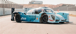 A race car designed for hydrogen racing is shown on a racetrack. The car features a striking light blue and white design with bold graphics, sleek aerodynamic shapes, and large black wheels. The background includes a racetrack barrier and a soft-focus building in the distance.