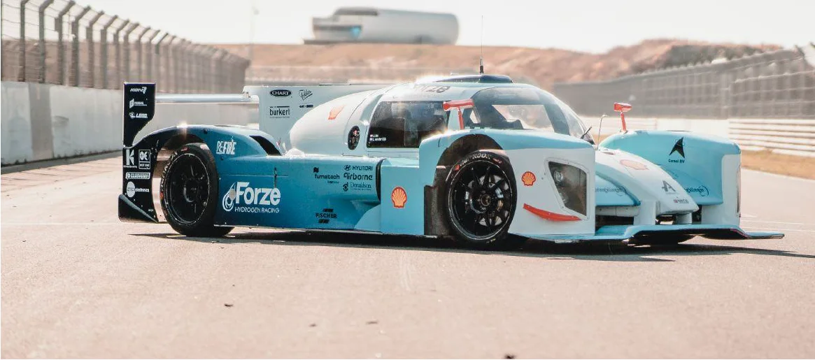 A race car designed for hydrogen racing is shown on a racetrack. The car features a striking light blue and white design with bold graphics, sleek aerodynamic shapes, and large black wheels. The background includes a racetrack barrier and a soft-focus building in the distance.