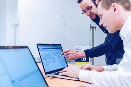 Two men are collaborating at a desk, focused on a laptop screen. One points to the screen while the other types. A box labeled "The Emotional Experts" is visible in the background. The setting appears to be a modern office.