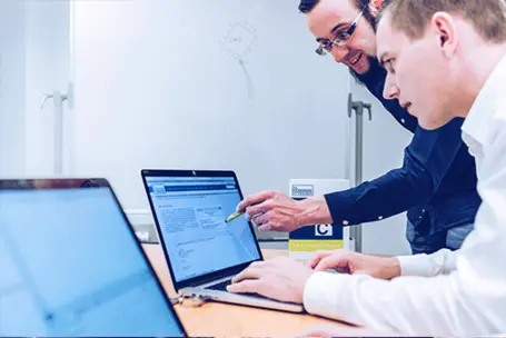 Two men are collaborating at a desk, focused on a laptop screen. One points to the screen while the other types. A box labeled "The Emotional Experts" is visible in the background. The setting appears to be a modern office.