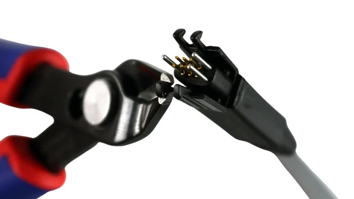 A close-up image of a pair of wire cutters gripping an electrical connector. The connector features several metal pins, some gold-plated, visible inside. The background is plain white, emphasizing the tools and connection.