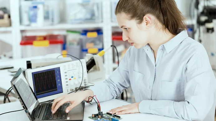 A woman is focused on her work at a laboratory table. She is using a laptop connected to a circuit board, with an oscilloscope displaying waveforms nearby. The setting is equipped with various electronic tools and materials.