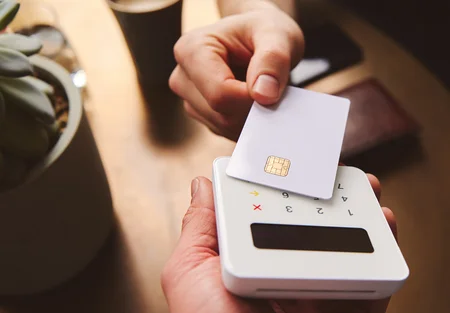 A person is using a card reader, holding a blank card above it. The card reader has a numeric keypad and a screen, indicating a transaction. A plant is visible in the background, adding a casual ambiance to the scene.