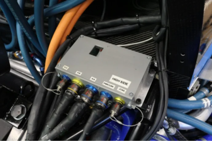 A rectangular gray control unit labeled "HIGH SIDE" with multiple colored connectors is situated among a tangle of blue, orange, and black hoses. This setup likely indicates a complex mechanical or electrical system, possibly related to automotive or engineering technology.