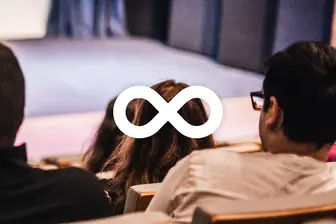 In a theater, the back of two seated individuals is visible. A symbol resembling an infinity sign is overlaid in the center, suggesting themes of continuity or limitless possibilities, amidst a blurred stage backdrop. The image conveys a sense of engagement in a performance or discussion.