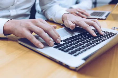 A close-up view of a person's hands typing on a laptop keyboard, set on a wooden table. Another laptop is visible in the background, suggesting a collaborative work environment. The focus is on the act of typing.