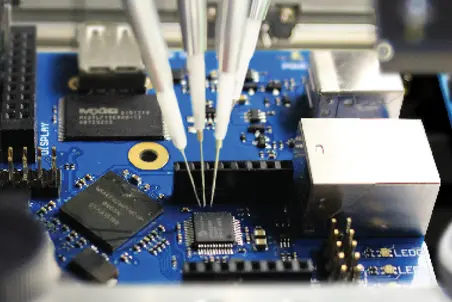 A close-up view of a blue circuit board featuring various electronic components. Three thin probes are positioned over a small black microchip, indicating a testing or connection process. Additional components, including connectors and metallic parts, are visible in the surrounding area.