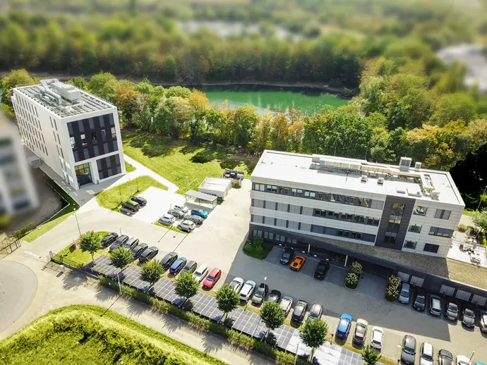 An aerial view of two modern buildings surrounded by green landscaping and a pond. A parking lot filled with cars is visible in front, with solar panels installed on one side. The environment features trees with changing fall colors.