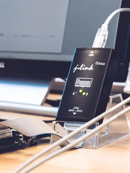 A black SEGGER J-Link Base debugging tool is prominently featured, connected by a USB cable. It has indicator lights and is positioned on a clear stand, with a flat ribbon cable connected to it. A blurred computer monitor is visible in the background.