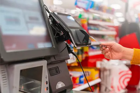 A close-up of a self-checkout kiosk in a retail store. A person's hand, holding a payment card, is positioned near a card reader, ready to process a transaction. The background features shelves stocked with various products.