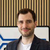 A smiling man stands in front of a modern background featuring wooden panels and a logo with a blue arrow. He is wearing a blazer over a white t-shirt. Shelves behind him display various tech products.
