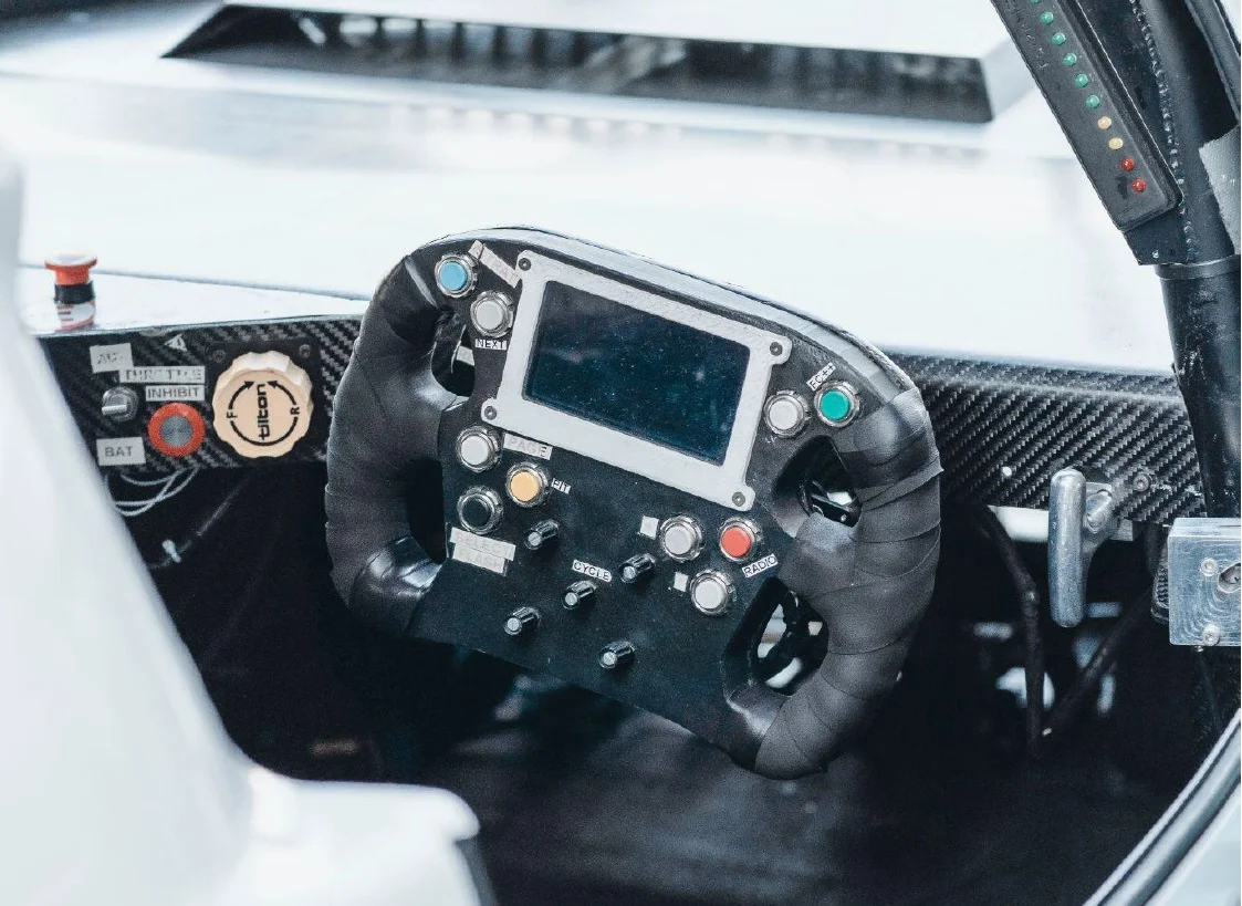 A close-up view of a racing car steering wheel, featuring various buttons and a digital display. The wheel is wrapped in black material for grip, and there are several colorful buttons for controls, along with a carbon fiber dashboard area.