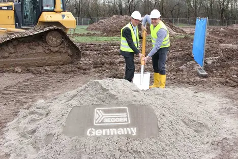 Three construction workers in safety vests and hard hats stand around a mound of sand, preparing to break ground for a project. The mound features a logo and the word "Germany" etched into the surface, indicating the start of construction for a facility. A bulldozer is in the background.